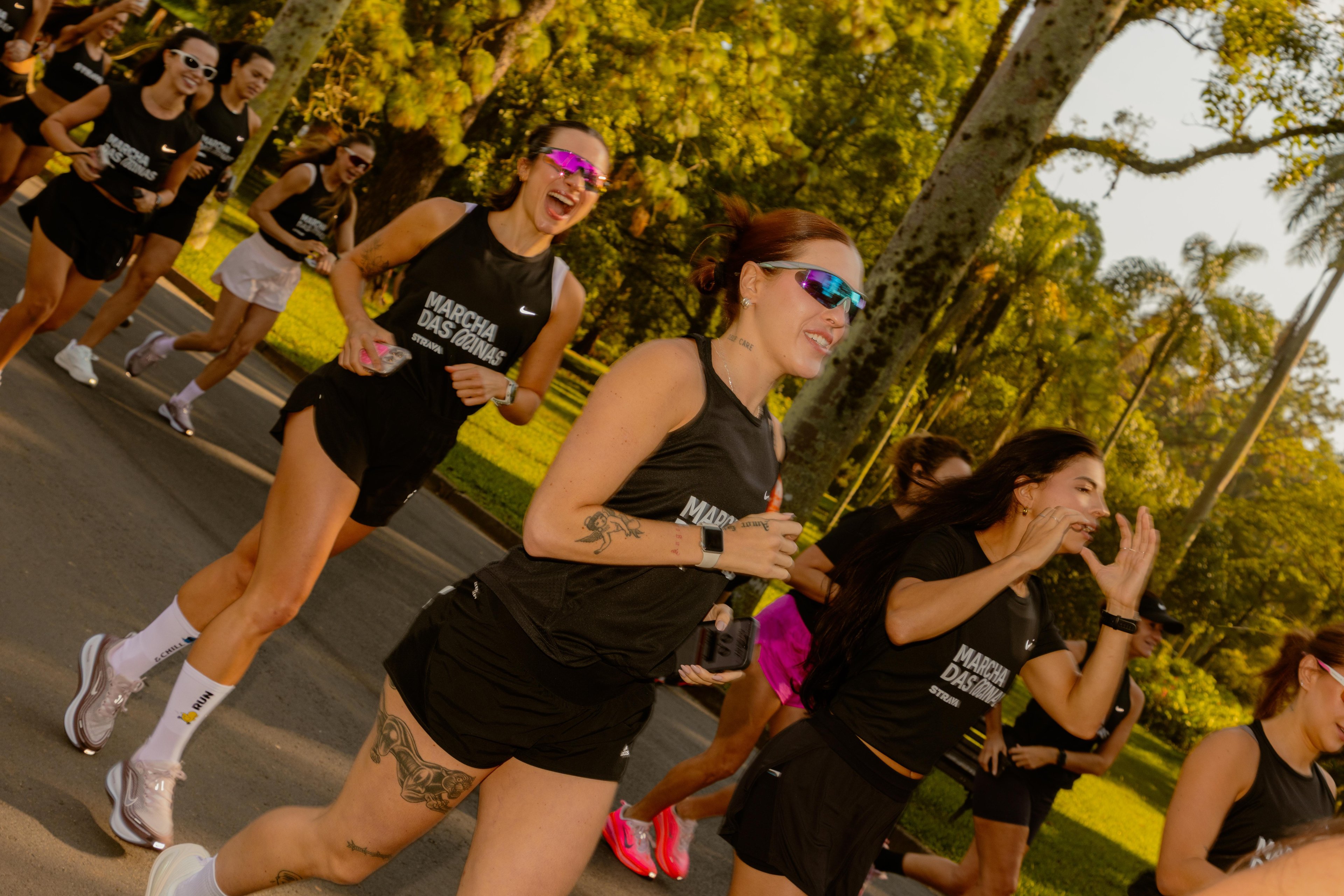 Corredoras strava em regatas e shorts pretos correm sorrindo no parque ao sol dourado. Texto: MARCHA DAS OCINAS STRAVA.