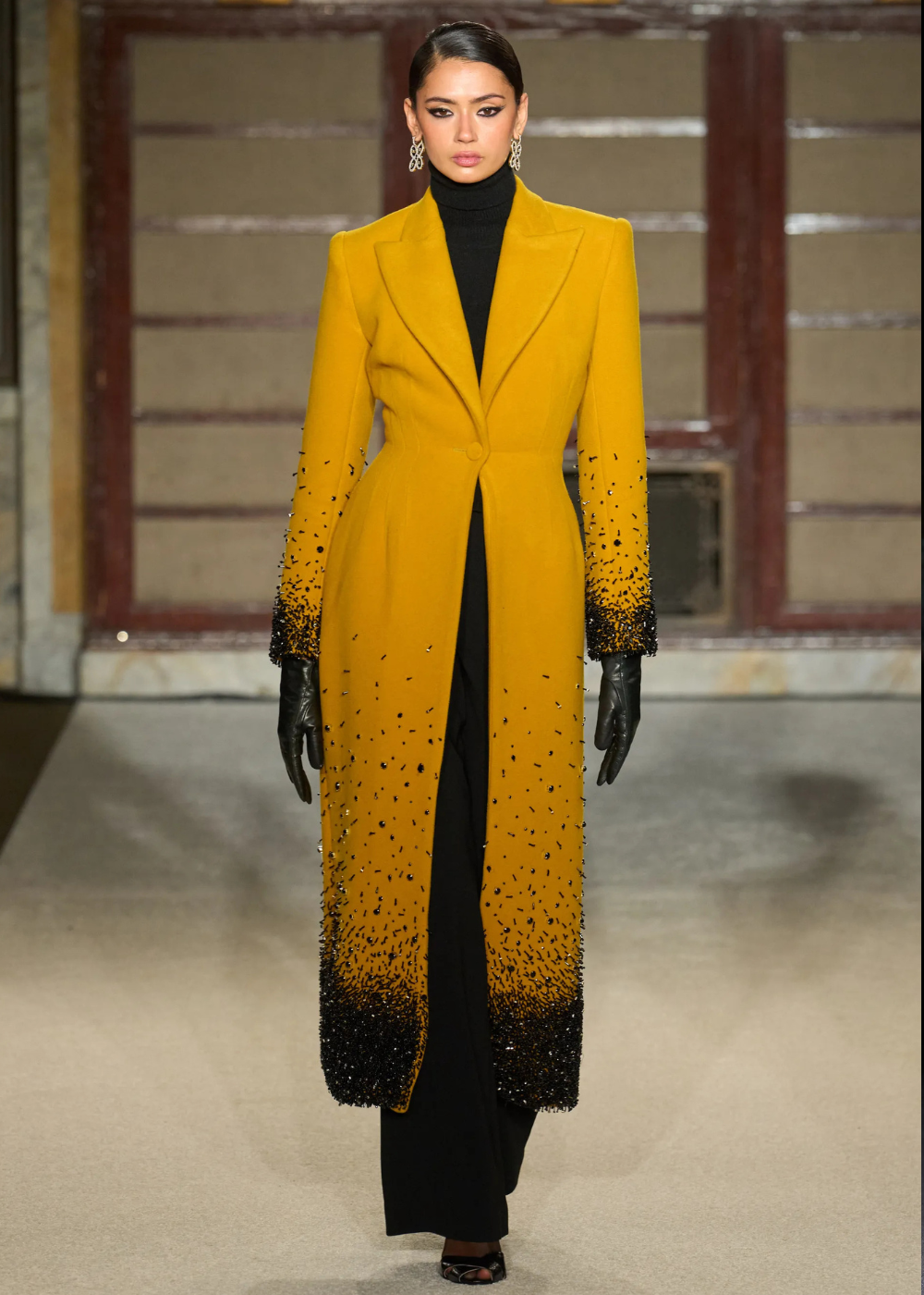 Outono/Inverno 2026: modelo posa com casaco amarelo acinturado, gola alta preta, luvas de couro e brilho preto, clima