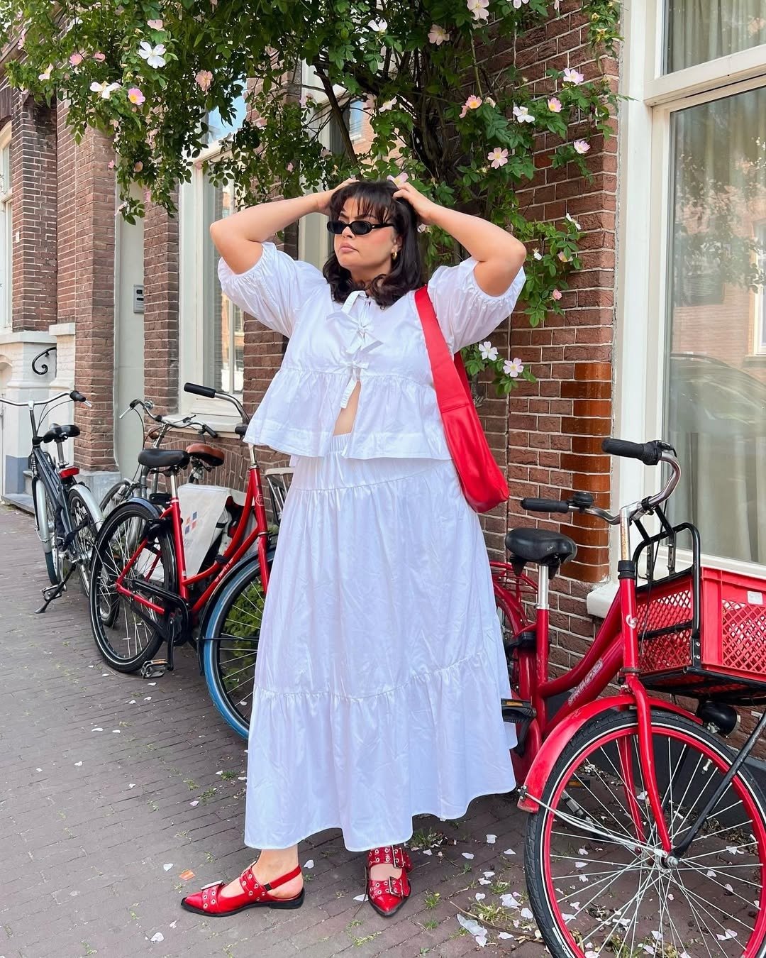 Modelo posa com bolsa vermelha, vestido branco amplo de algodão e sapatilhas vermelhas em rua florida, clima leve.