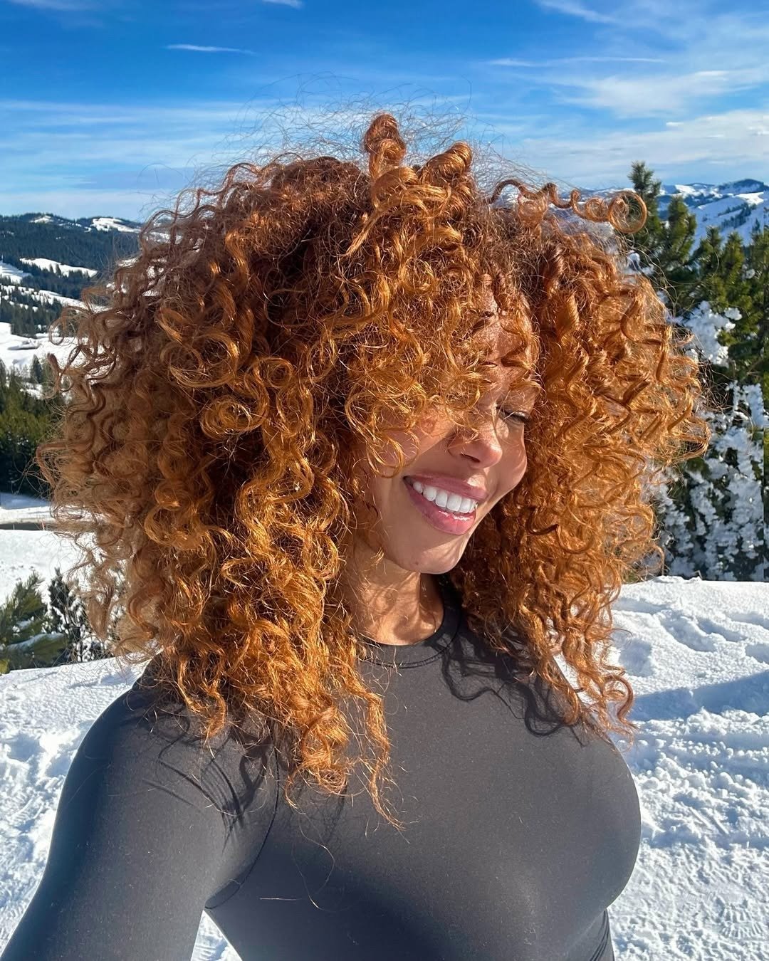Opções de tinta para cabelo ruivo em cachos volumosos acobreados; modelo posa sorrindo na neve com blusa preta justa.