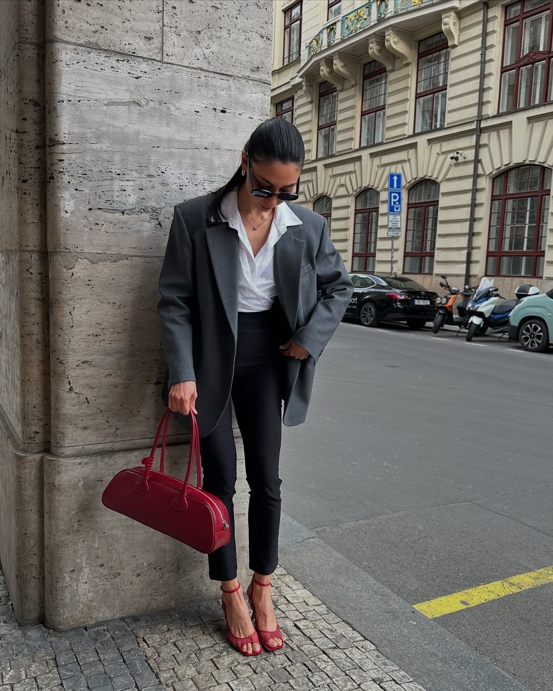 Modelo posa com bolsa vermelha de couro, blazer cinza oversized, camisa branca e salto vermelho em clima urbano elegante.