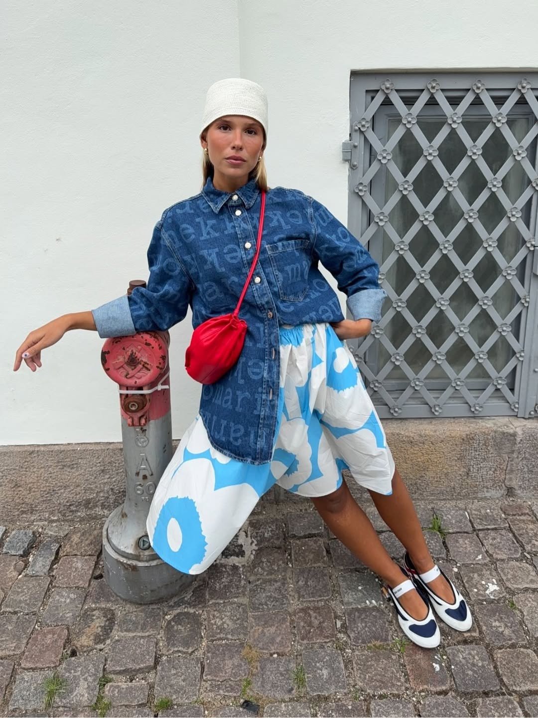 Modelo posa com bolsa vermelha, camisa jeans, saia branca e azul ampla e sapatilha bicolor em clima urbano leve.