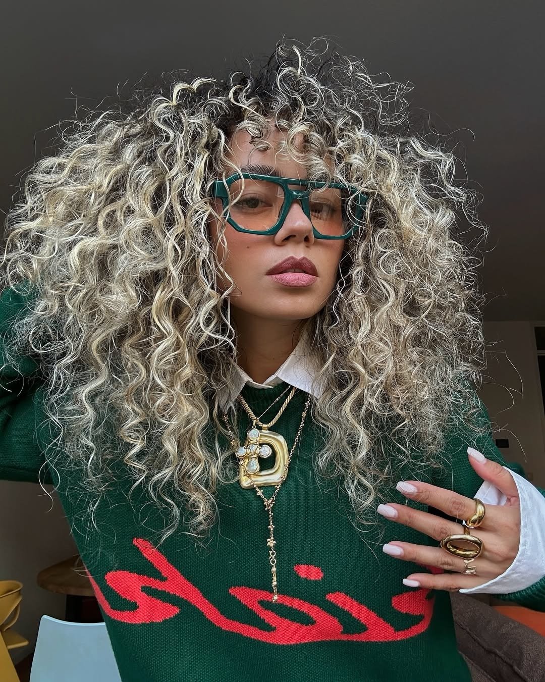Melhor shampoo para cabelos oleosos e finos inspira modelo com cachos volumosos, tricô verde e óculos marcantes.