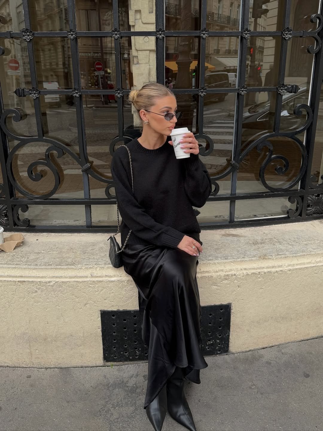 Looks com bota de bico fino: modelo posa sentada com tricô e saia acetinada pretos, mood urbano elegante.