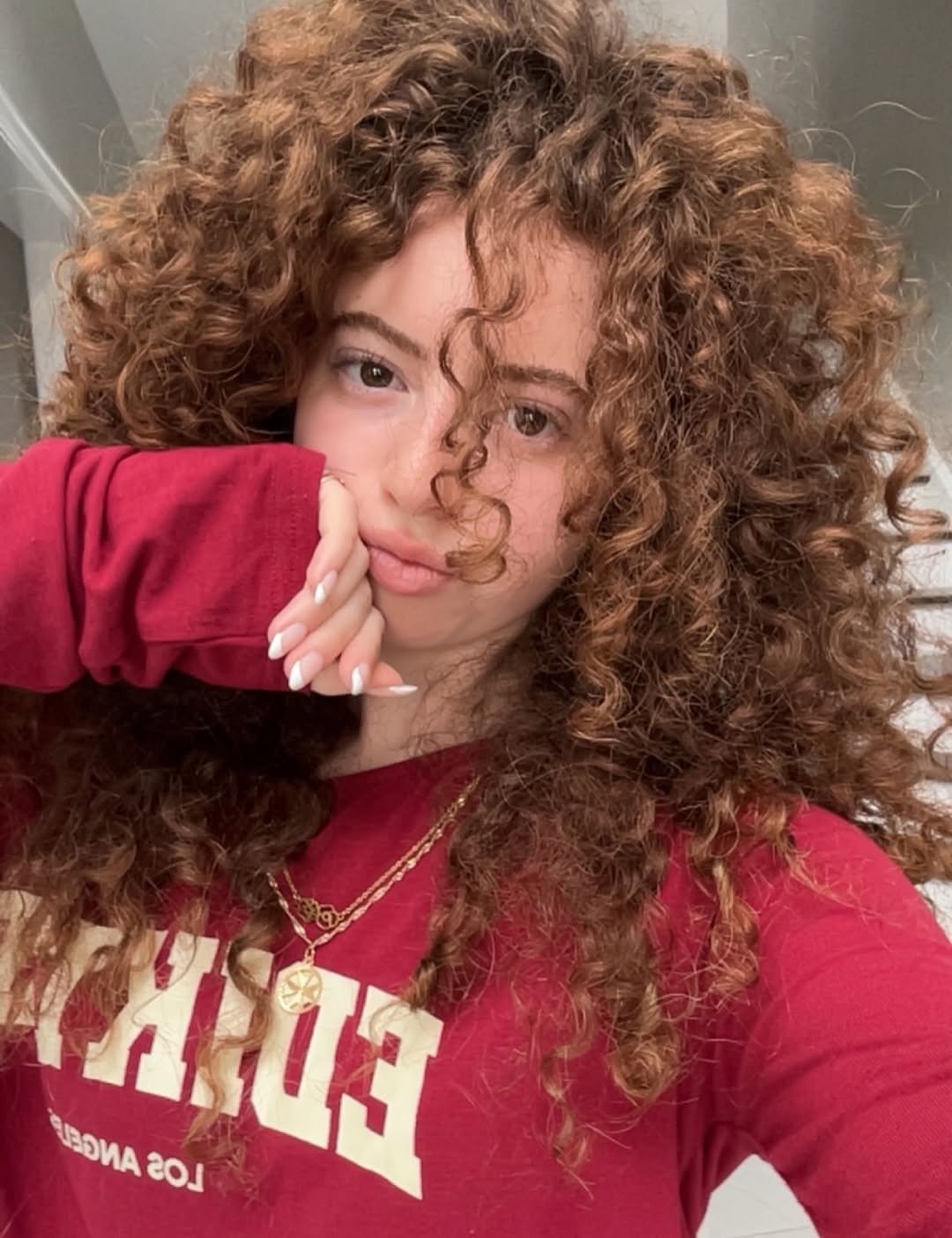Modelo posa com cabelo poroso cacheado castanho, moletom vermelho e colares dourados em clima casual.