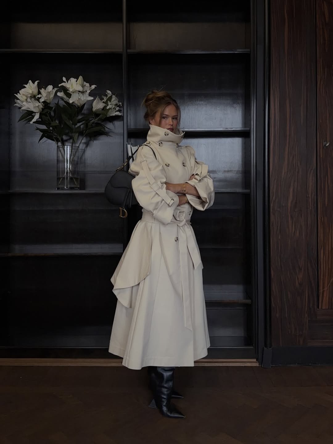 Modelo posa com trench coat bege acinturado, bolsa preta e looks com bota de bico fino em ambiente sóbrio e elegante.