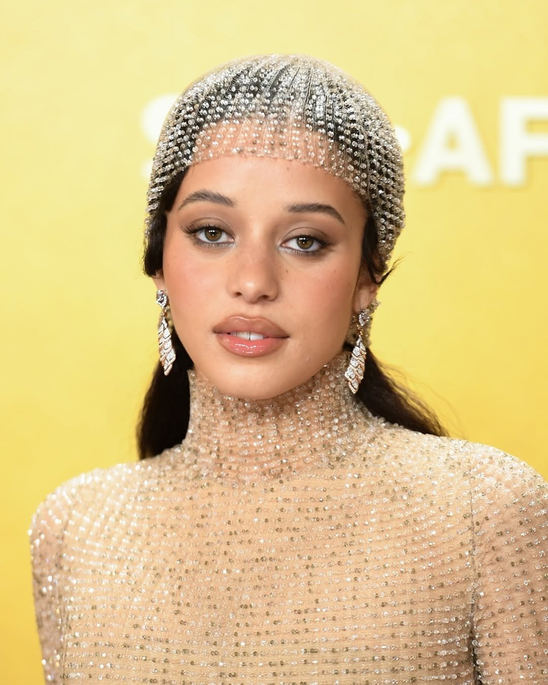 Modelo posa com halo lips em tom nude glossy, vestido e touca de malha translúcida cravejada de cristais, em fundo amarelo