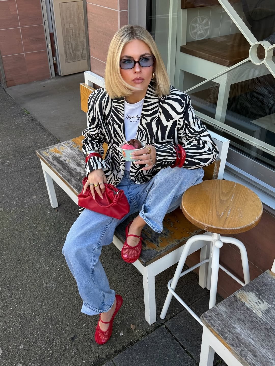 Modelo posa com bolsa vermelha, blazer zebra, jeans claros e sapatilha vermelha à mesa externa; Texto: Superdry est 2003