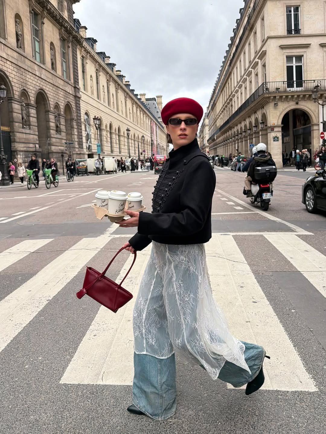 Modelo posa na faixa com bolsa vermelha, boina vinho, casaco preto e renda sobre jeans; clima urbano sofisticado.