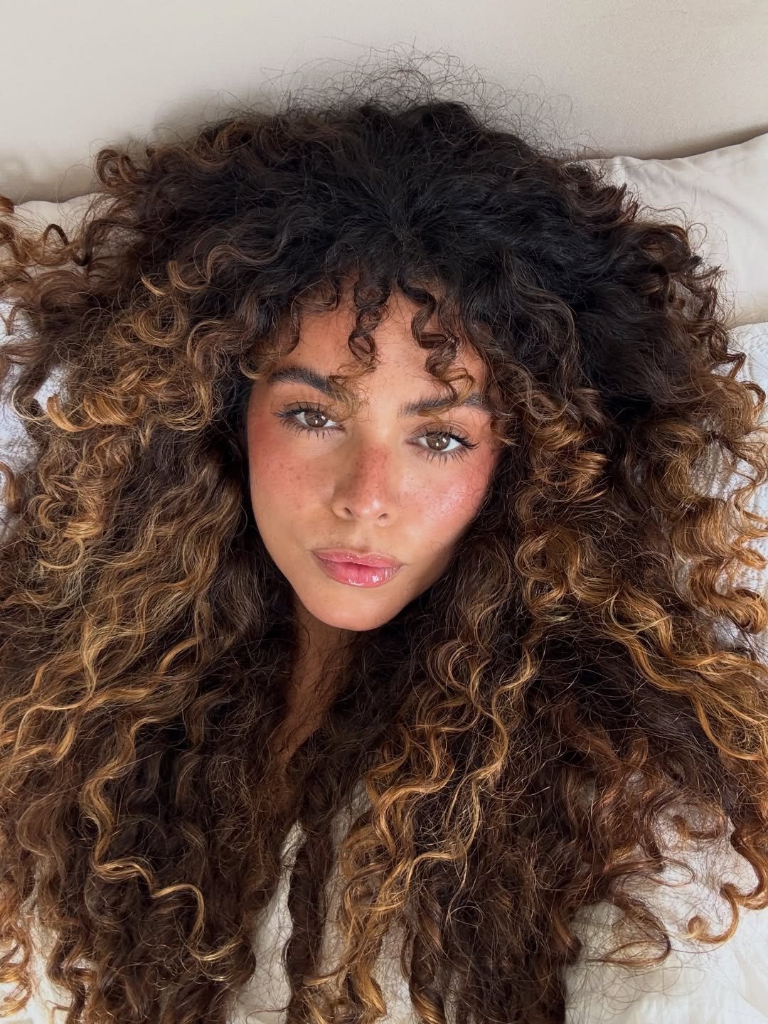 Modelo deitada exibe cachos volumosos castanho e mel em close suave, ideal para melhor shampoo para cabelos oleosos e finos.