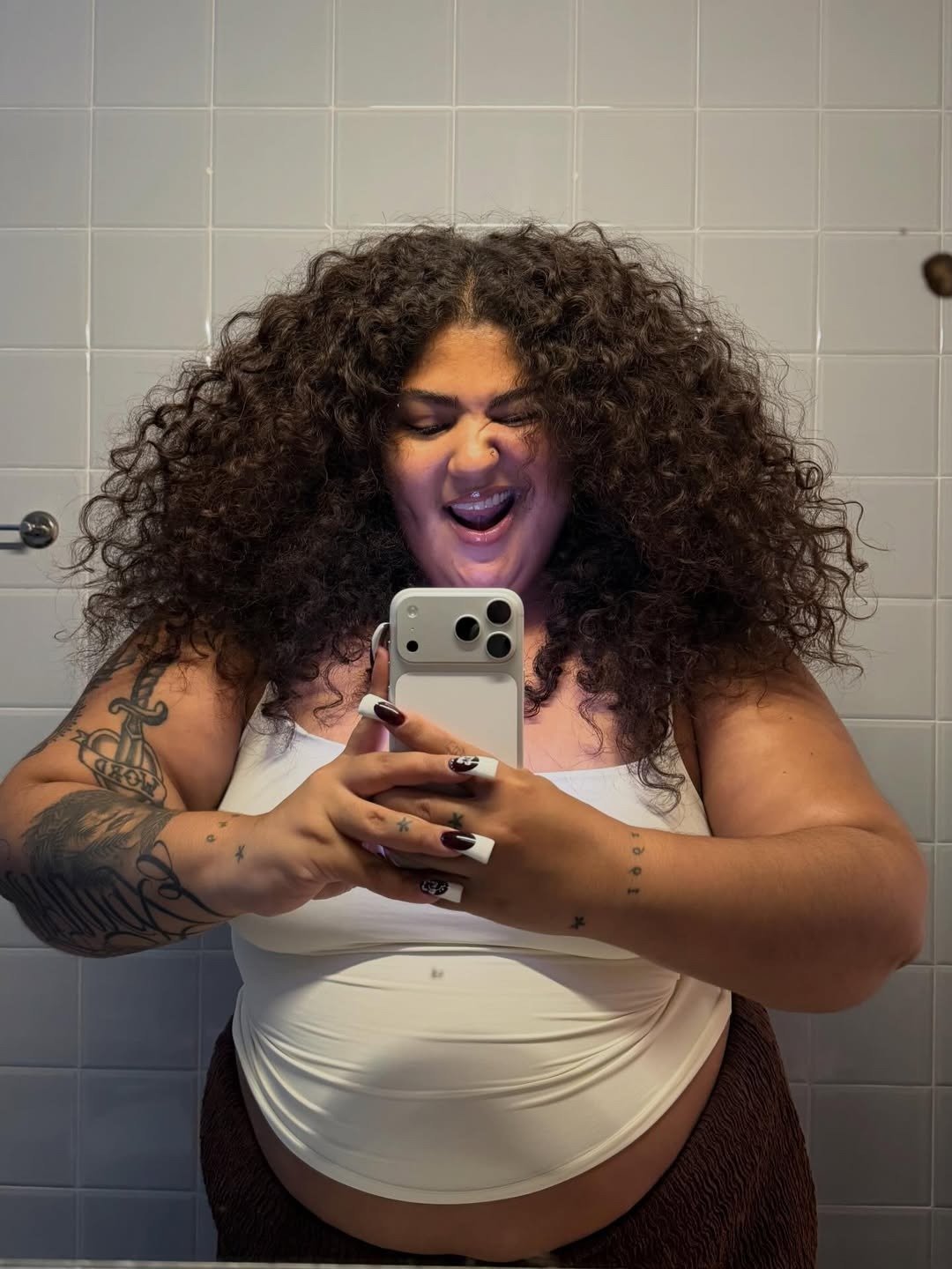 Modelo posa no espelho com top branco e cachos volumosos em banheiro bege, clima leve;