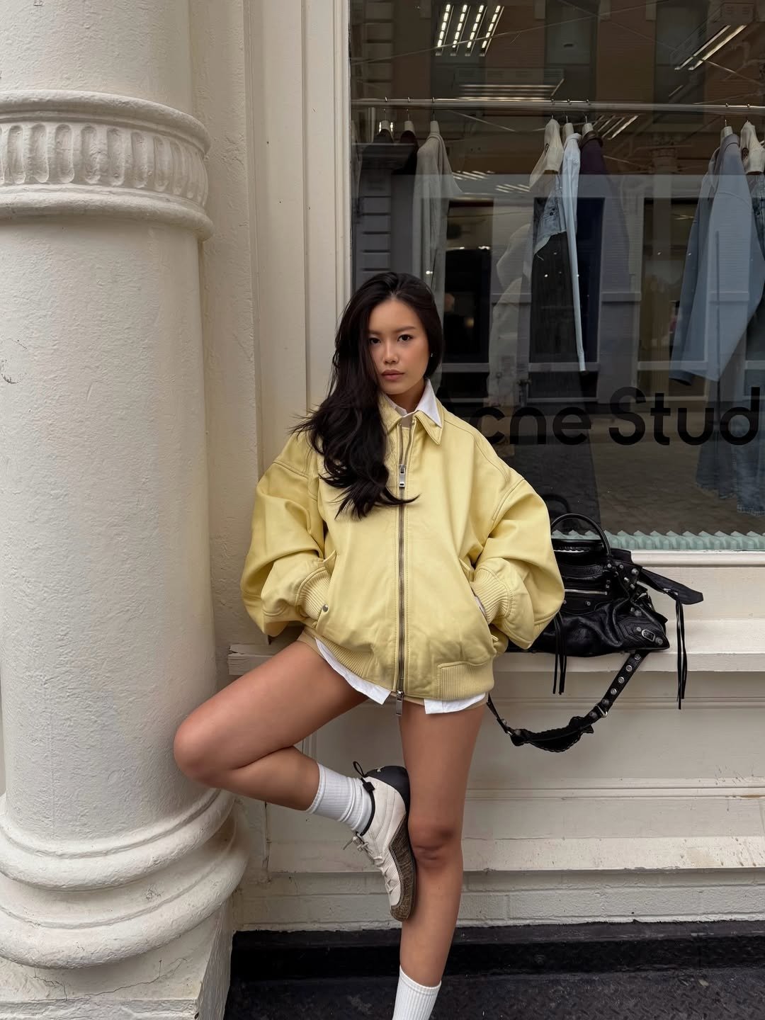 Modelo posa de jaqueta bomber amarela oversized e tênis branco diante de vitrine urbana; Texto: acne stuഡിയോ