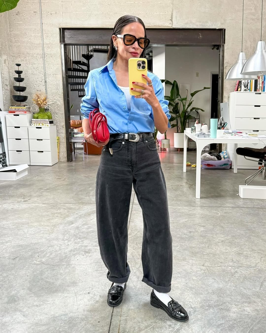 Modelo posa de mocassim feminino com calça jeans ampla preta, camisa azul e bolsa vermelha em ambiente moderno e elegante.