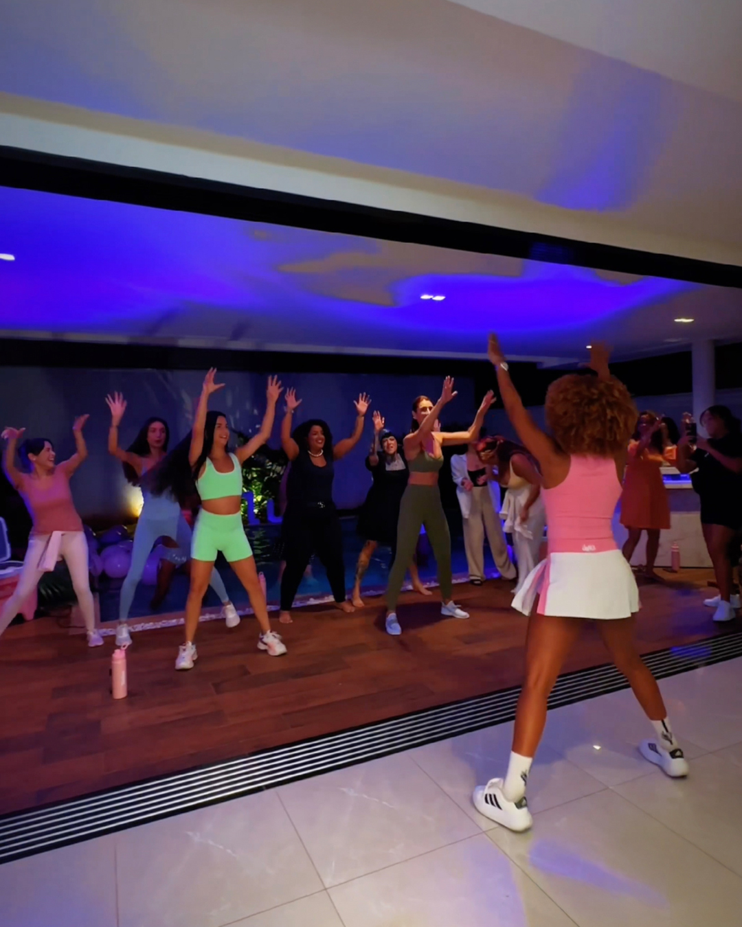 Marketing de experiência em aula fitness; instrutora de top rosa e saia branca conduz grupo animado sob luz azul e roxa.