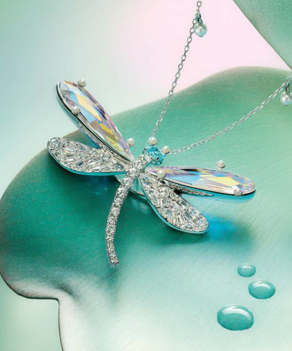 Colar Swarovski de libélula prateada com cristais iridescentes e pérolas sobre fundo verde-água, em clima delicado.