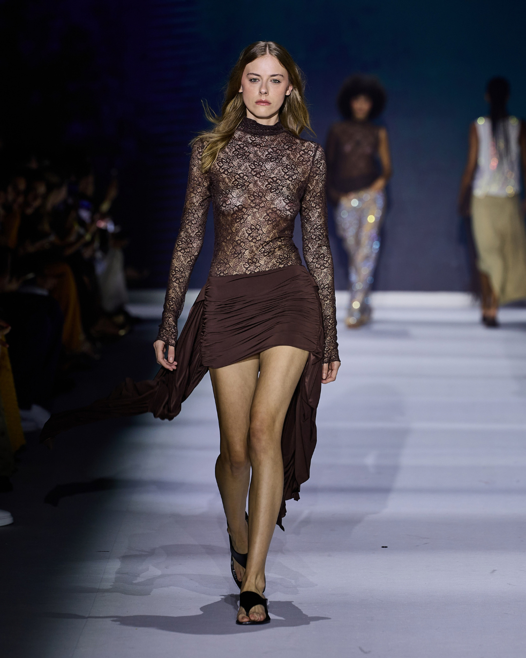 Tendências da primavera 2027: modelo desfila com renda marrom ajustada e saia drapeada assimétrica em clima sensual.