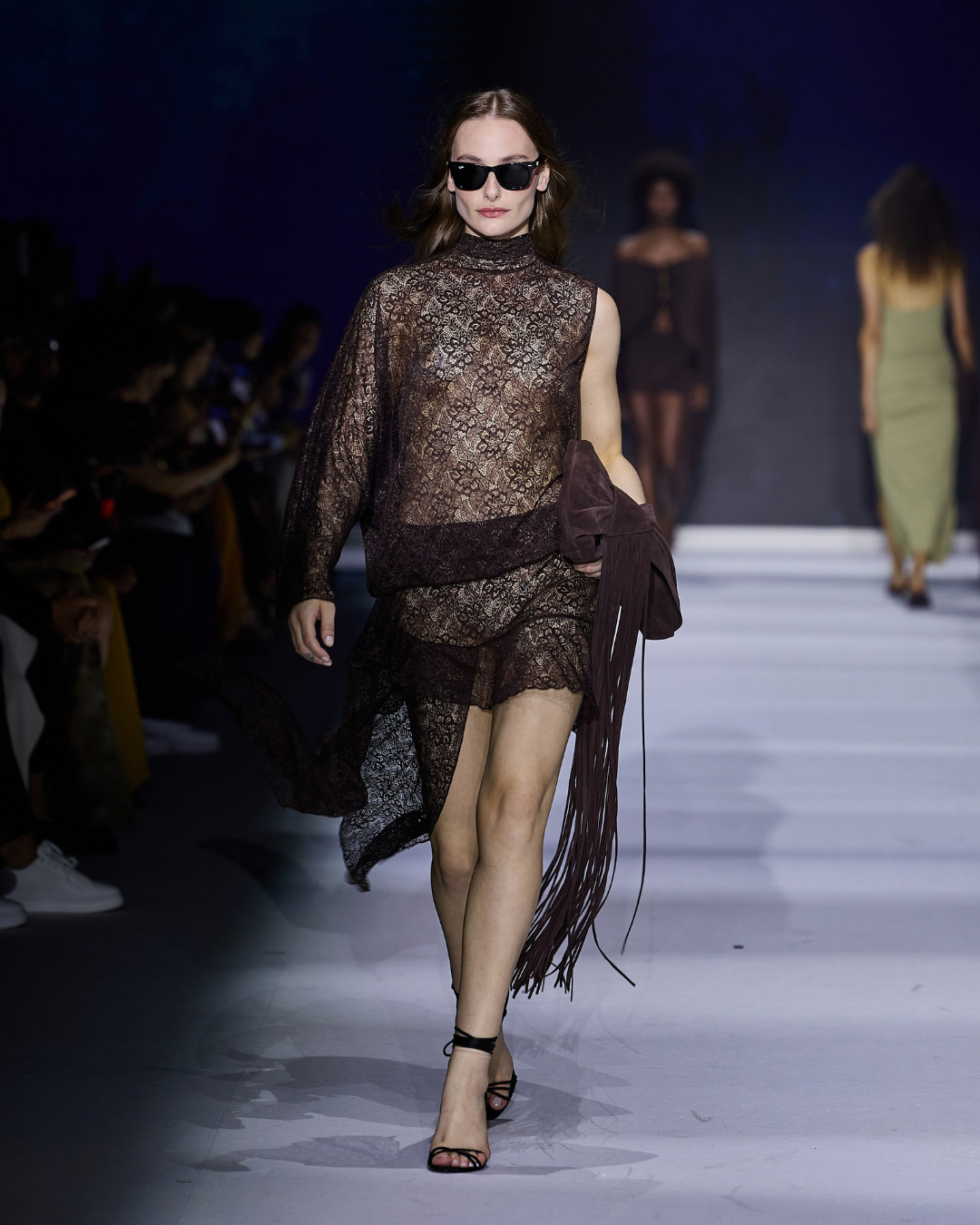 Modelo  com vestido assimétrico de renda marrom translúcida, bolsa de franjas e sandália preta - tendências de moda 2025