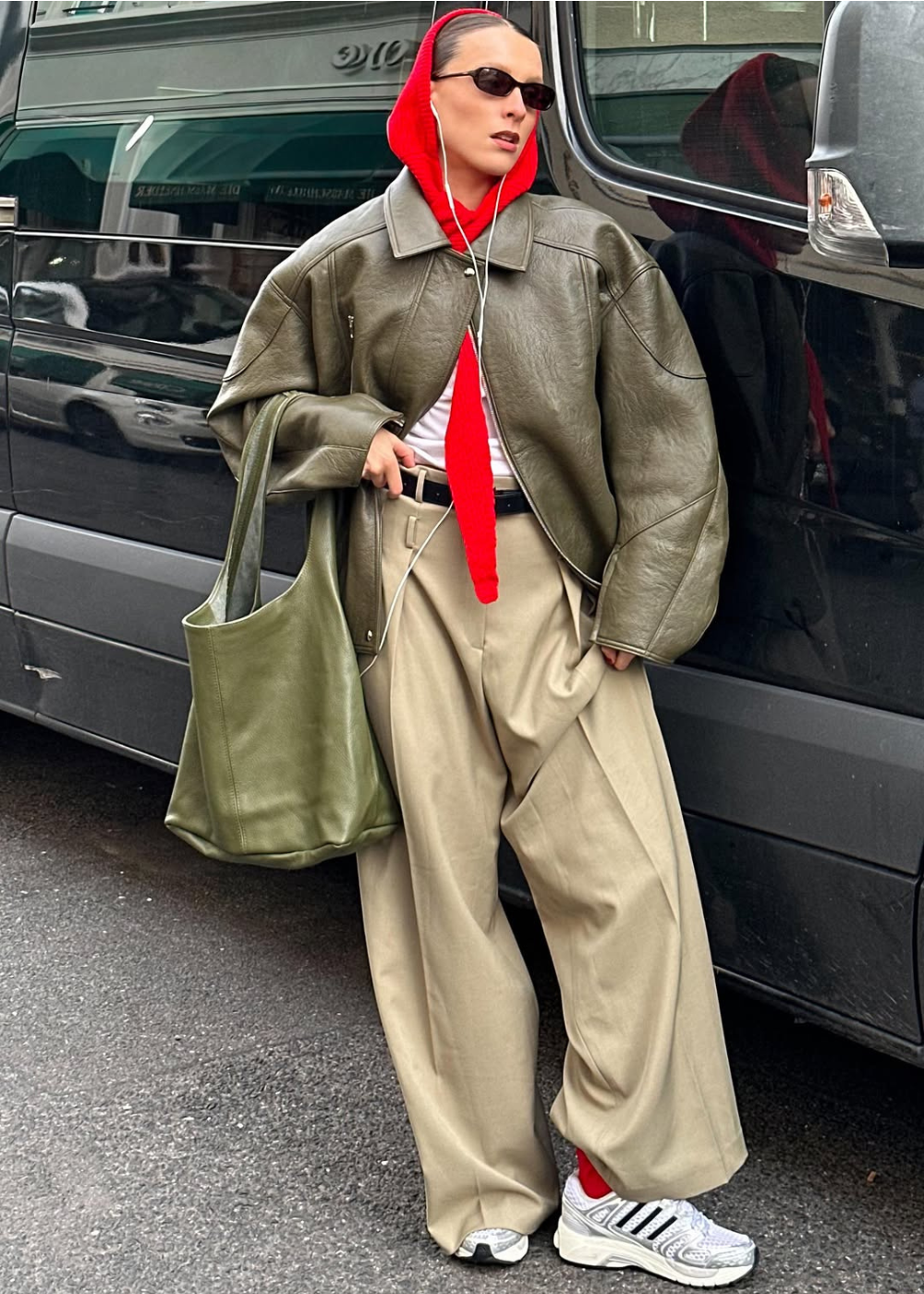 Modelo posa com calça pantalona bege e tênis esportivo, jaqueta verde oversized e capuz vermelho em clima urbano ousado.