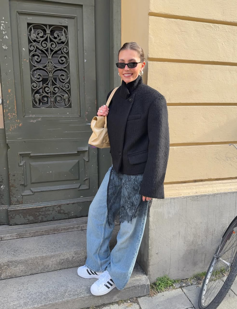Modelo posa com tênis clássico estiloso, jaqueta preta texturizada, jeans amplo e bolsa bege diante de fachada urbana.