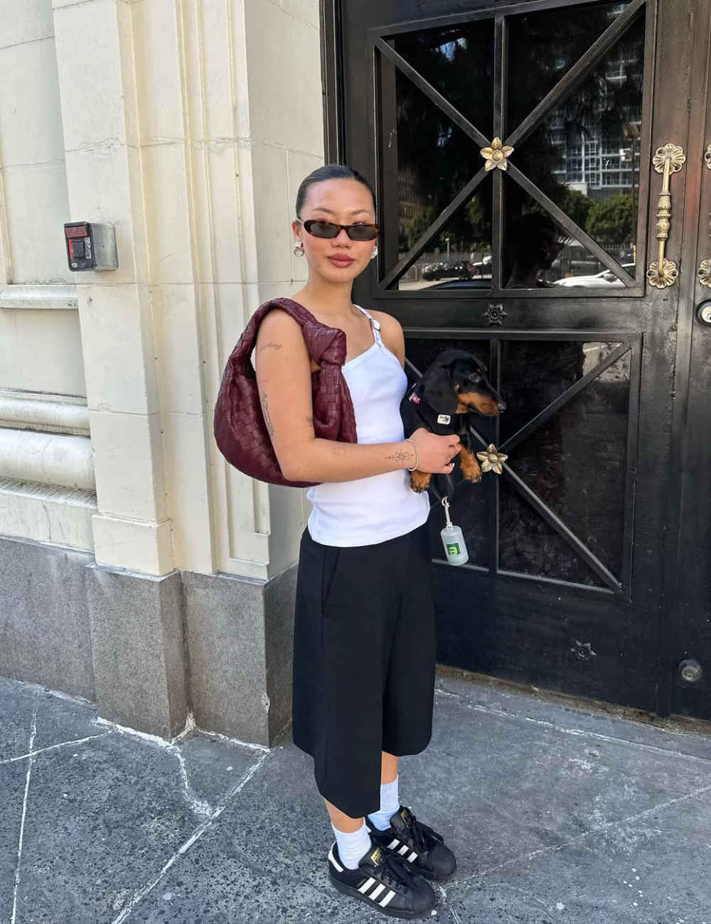 Modelo posa com tênis clássico estiloso preto e branco, regata branca, bermuda preta e bolsa vinho diante de porta elegante.