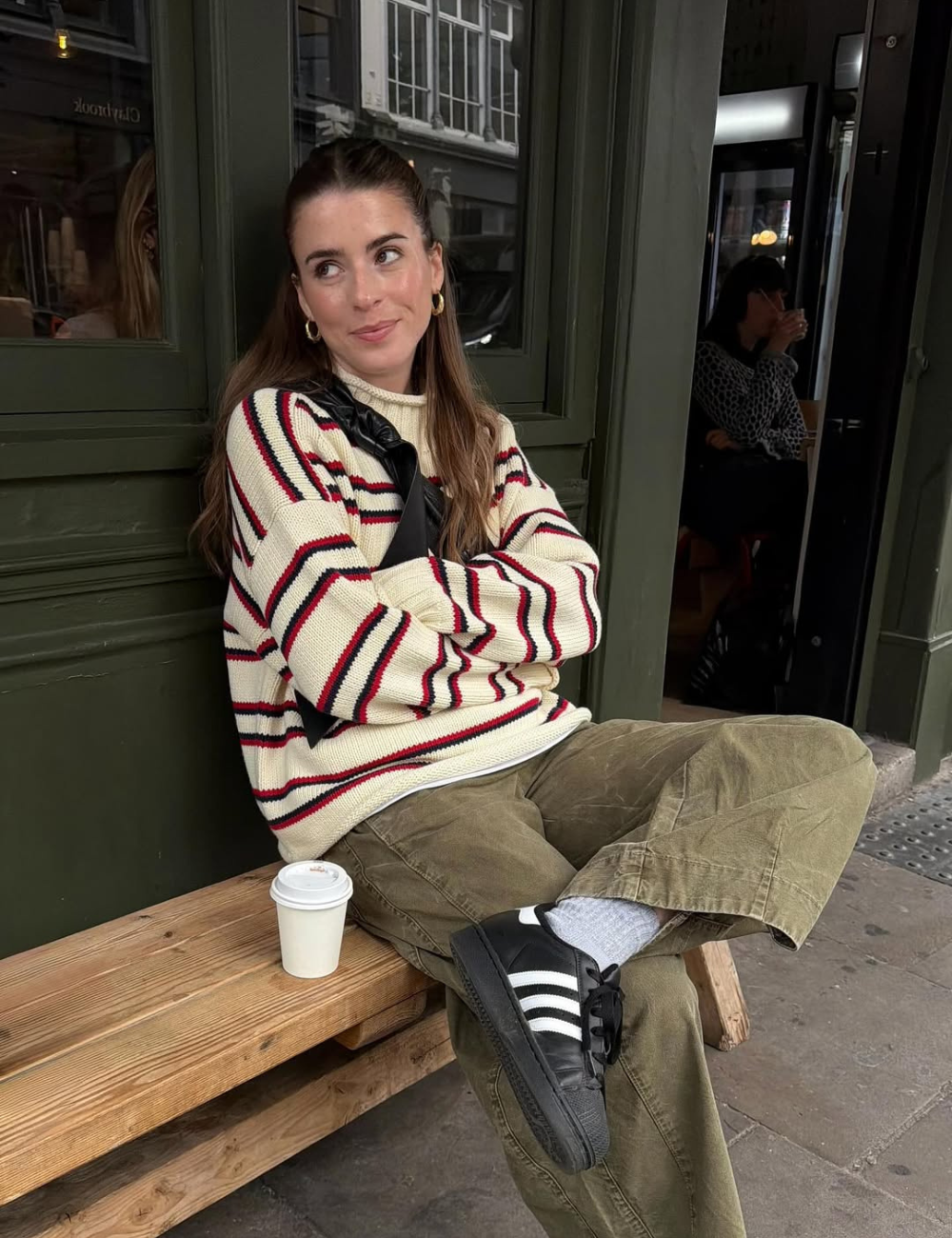 Modelo posa com tênis clássico estiloso preto e branco, tricô listrado e calça ampla verde em clima casual acolhedor.