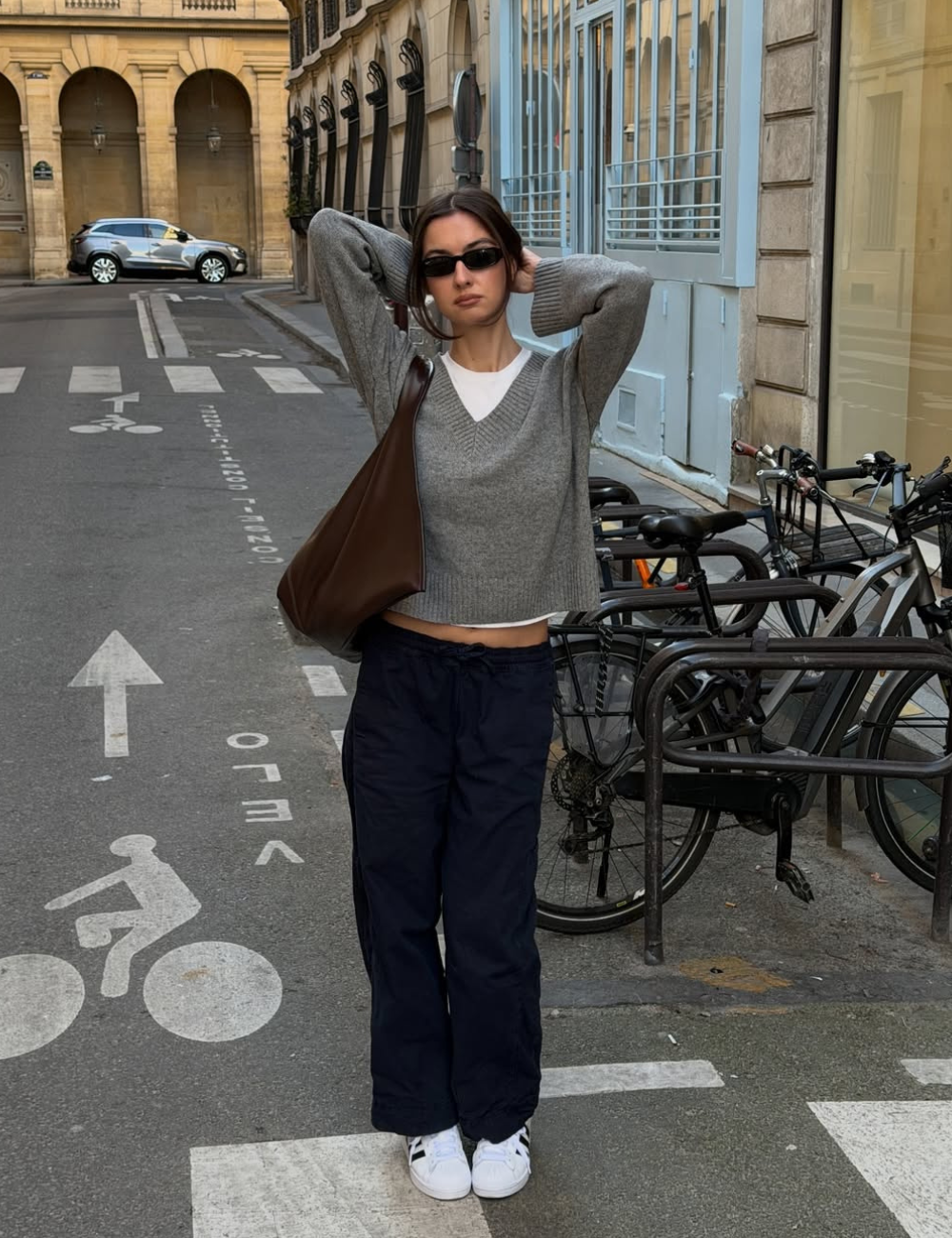 Modelo posa com tênis clássico estiloso, tricô cinza, calça azul-marinho e bolsa marrom em rua urbana elegante.