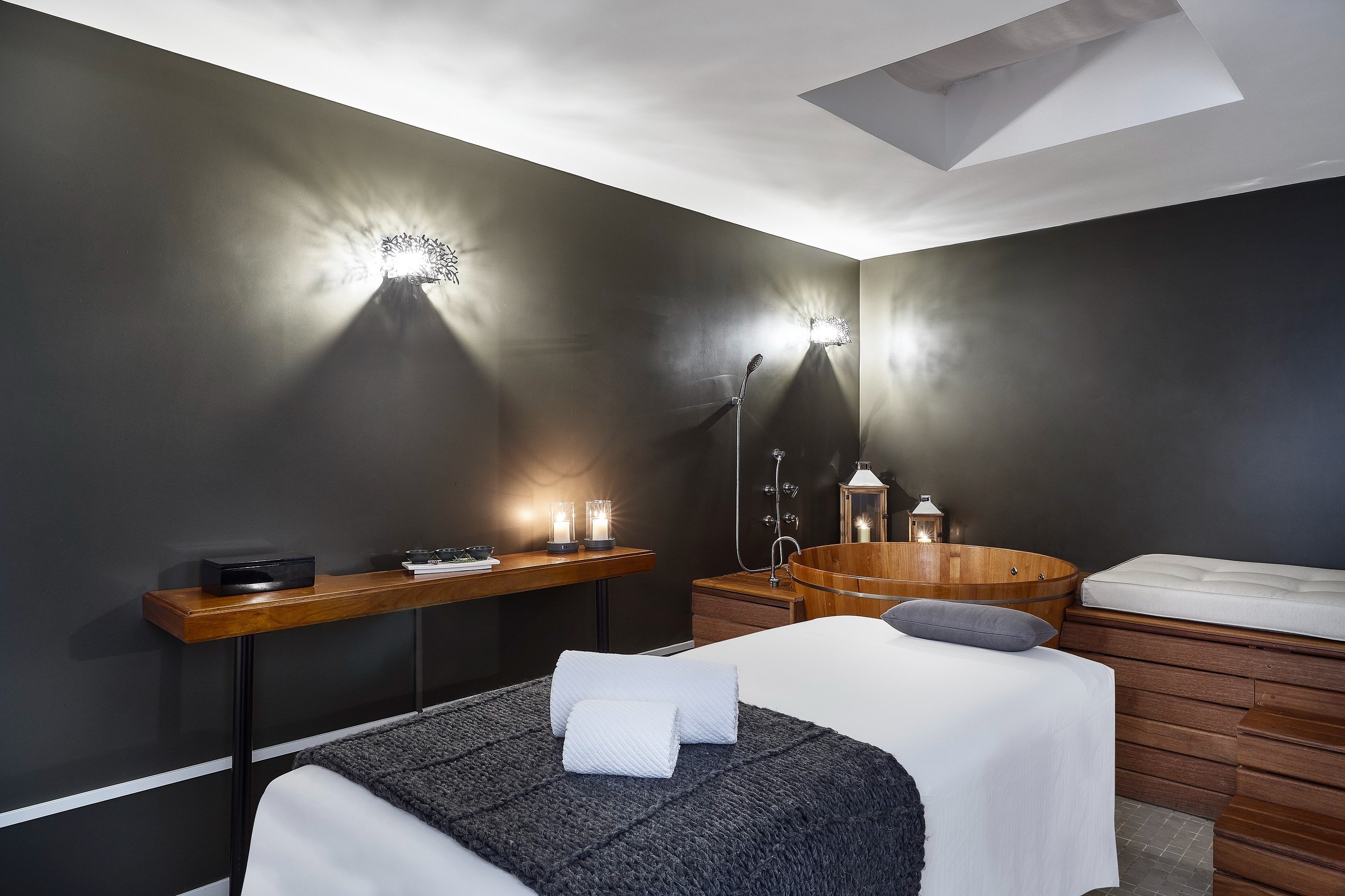 Sala de massagem com parede cinza escuro, mesa com toalha branca e manta cinza, banheira de madeira e iluminação suave com