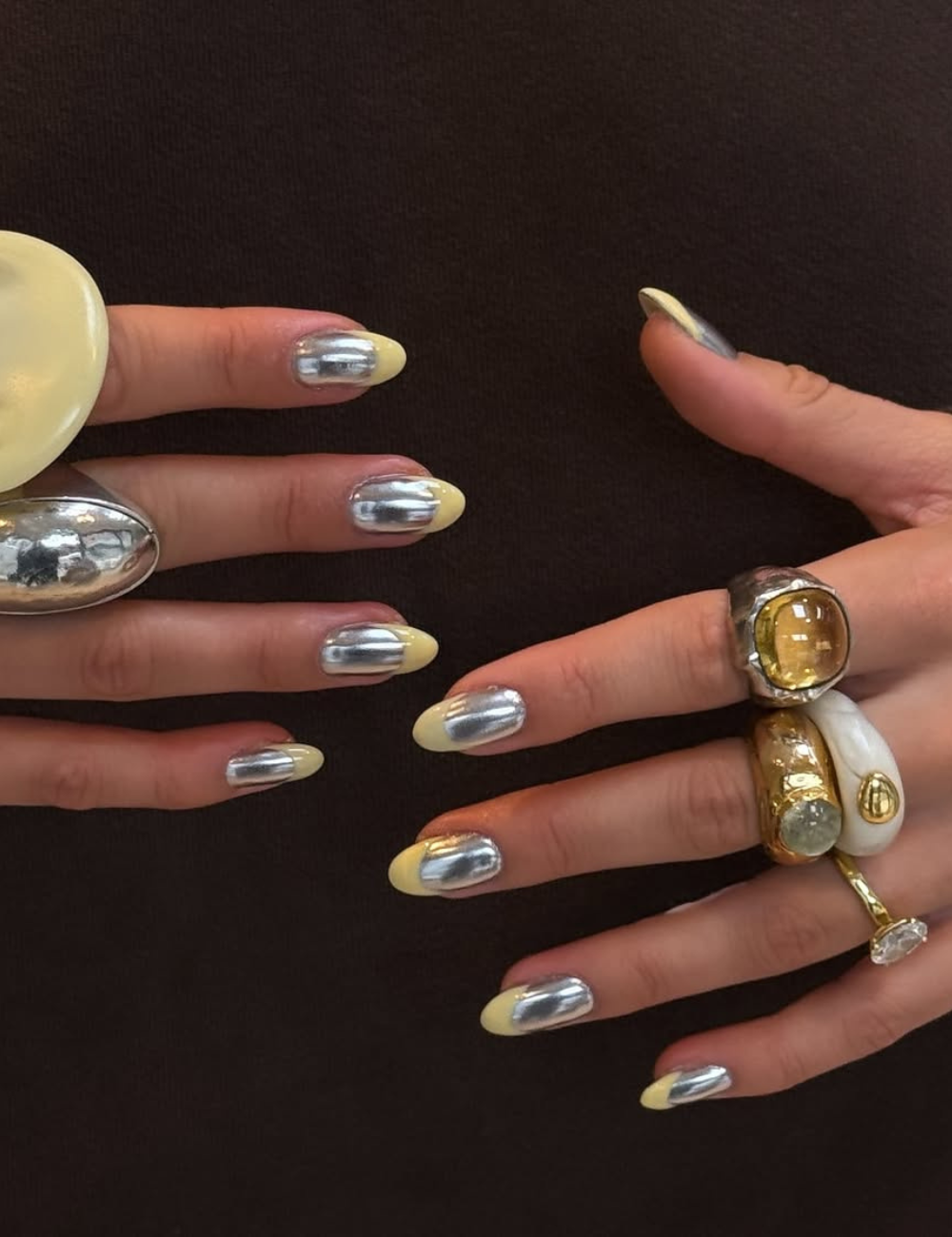 Unhas cromadas prateadas com pontas amarelo-claro e anéis esculturais dourados e brancos sobre fundo escuro elegante.