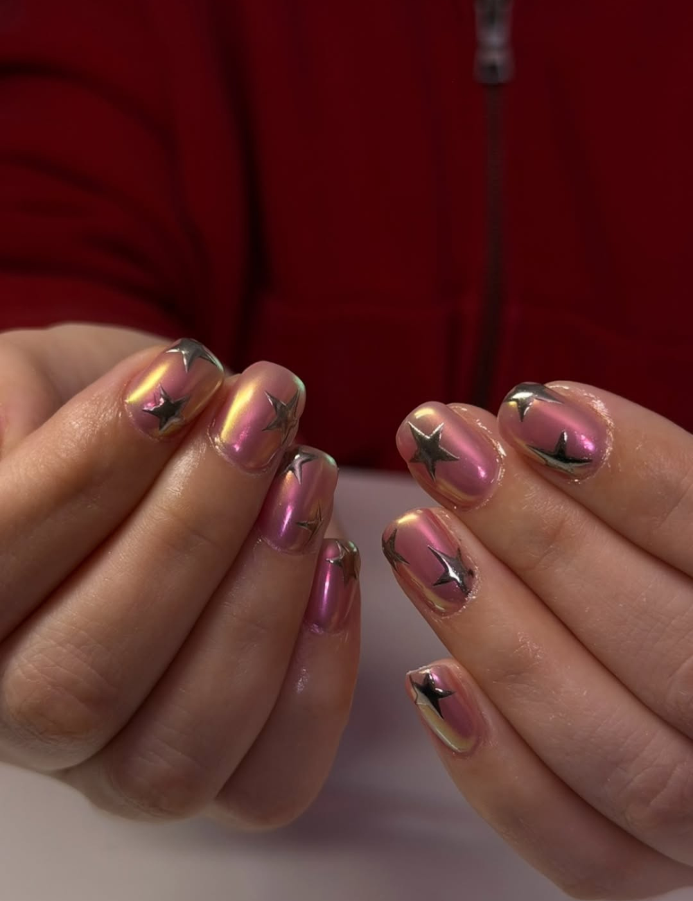 Unhas cromadas rosa furta-cor com estrelas prateadas ganham destaque nas mãos em pose delicada diante de blusa vermelha.