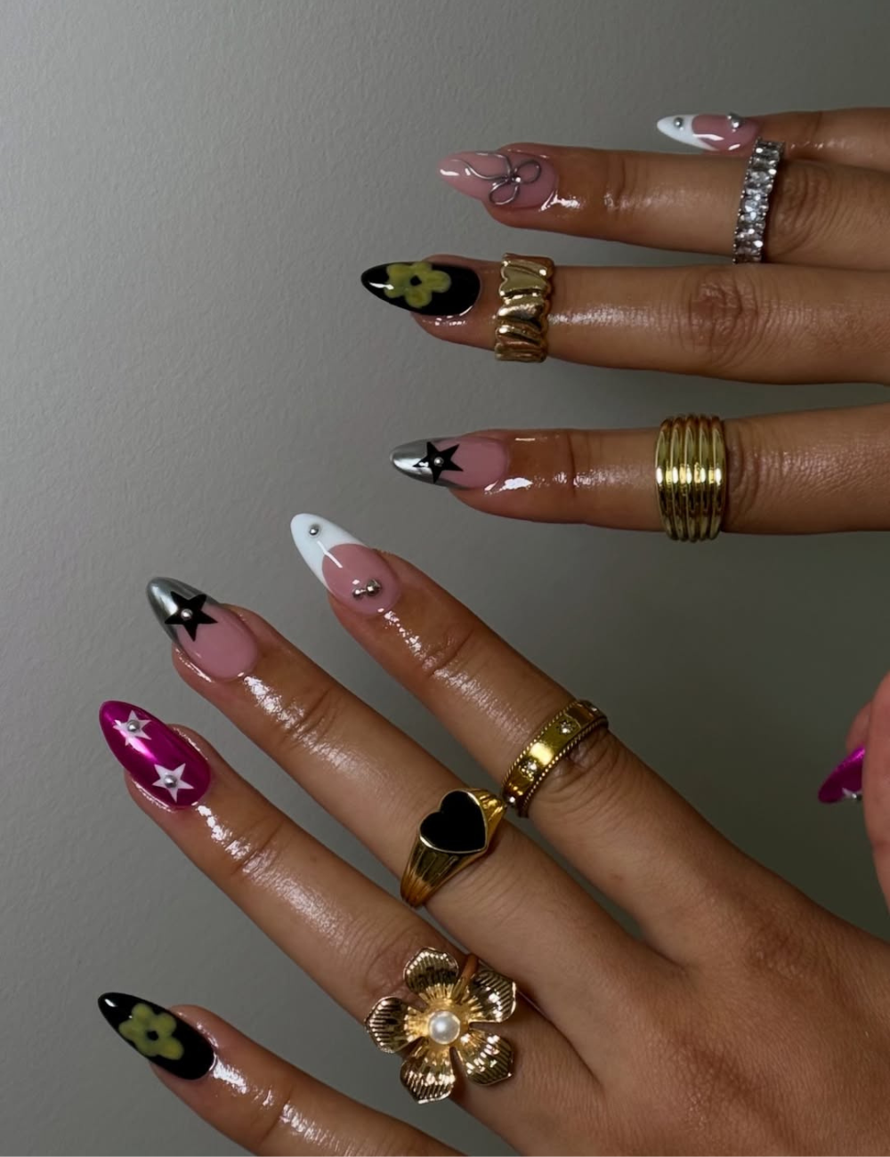Unhas cromadas e decoradas em rosa, prata, preto e fúcsia brilham com anéis dourados sobre fundo cinza.