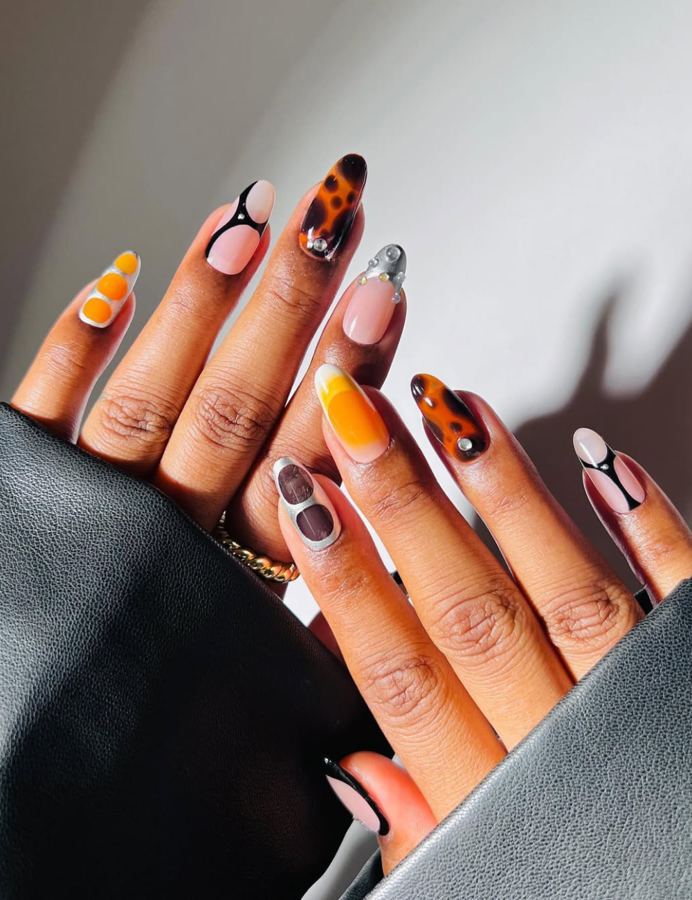 Unhas cromadas e nail art tartaruga, laranja e nude brilham em mãos posadas com mangas pretas, clima sofisticado.