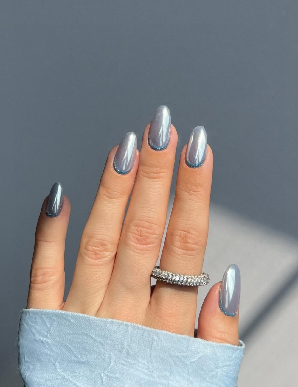 Unhas cromadas azul-prateadas em formato amendoado brilham sob luz suave, com anel cravejado e manga acetinada azul.