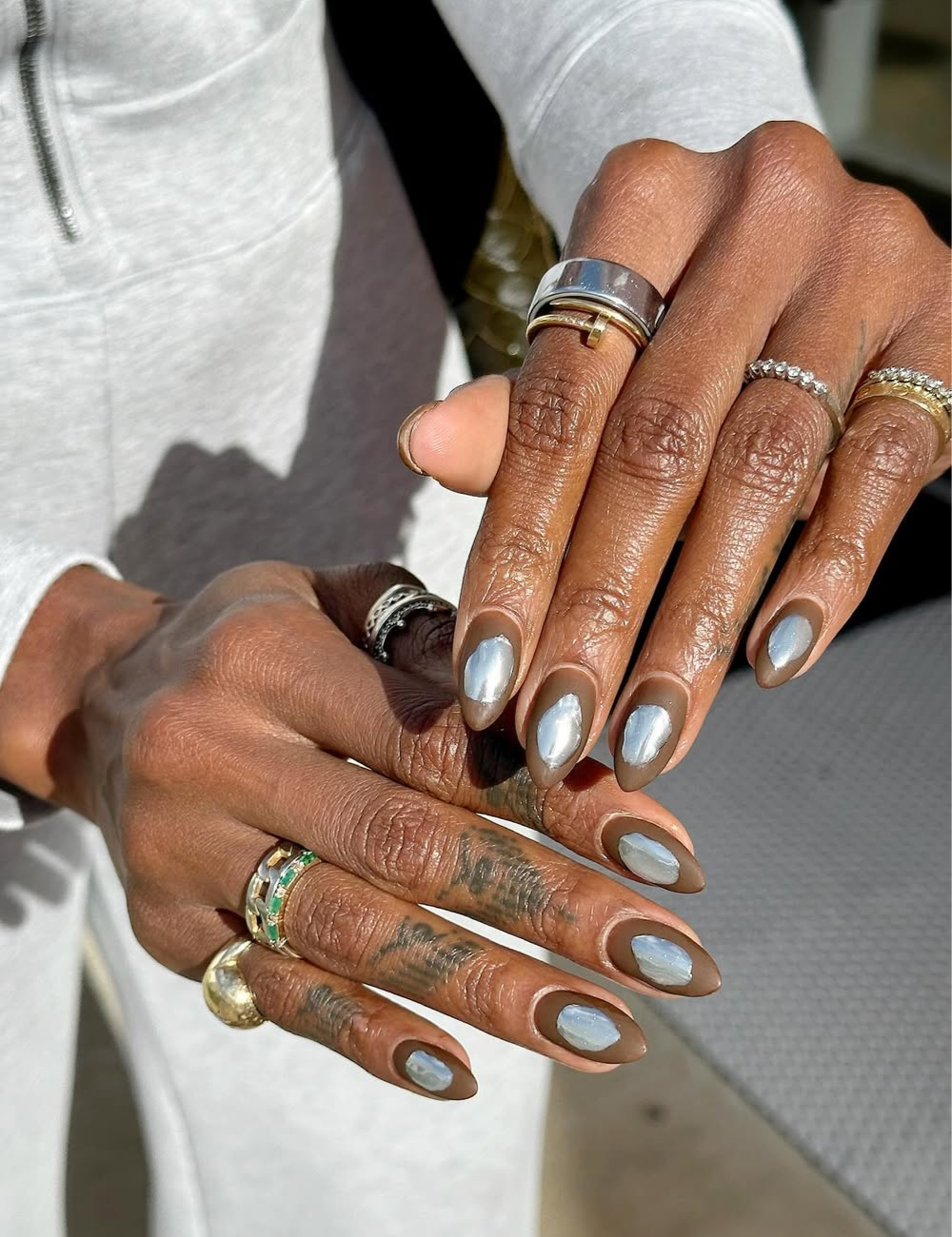 Unhas cromadas prateadas com contorno taupe brilham nas mãos com anéis, sobre look branco de malha em clima sofisticado.