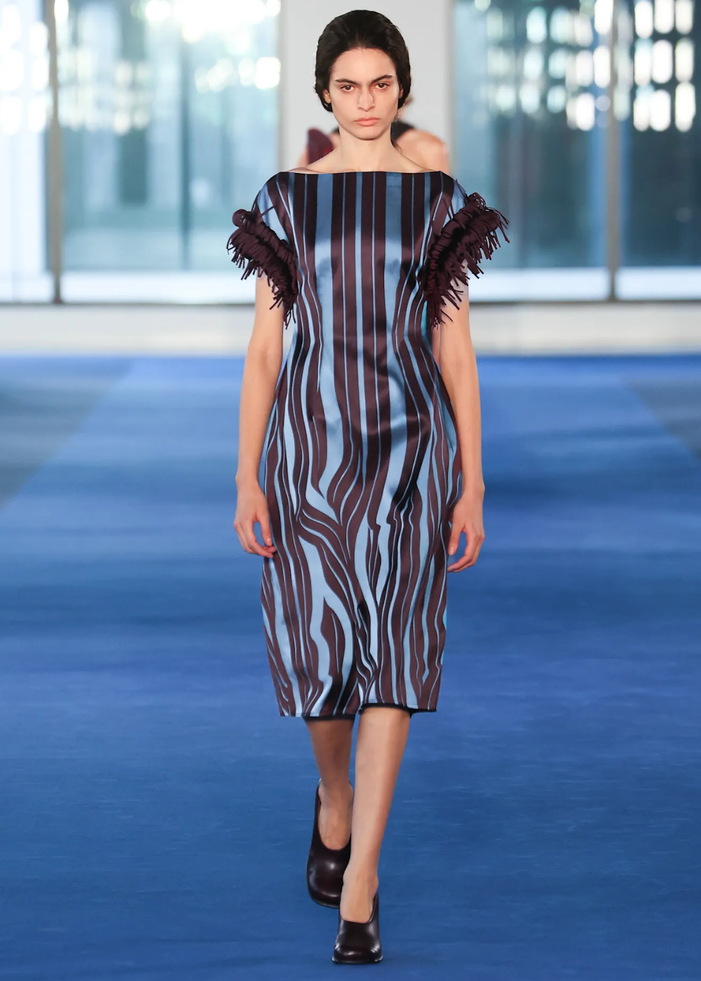 Modelo caminha com vestido midi azul e marrom listrado em tecido acetinado, mangas com franjas e vamp pumps marrons.