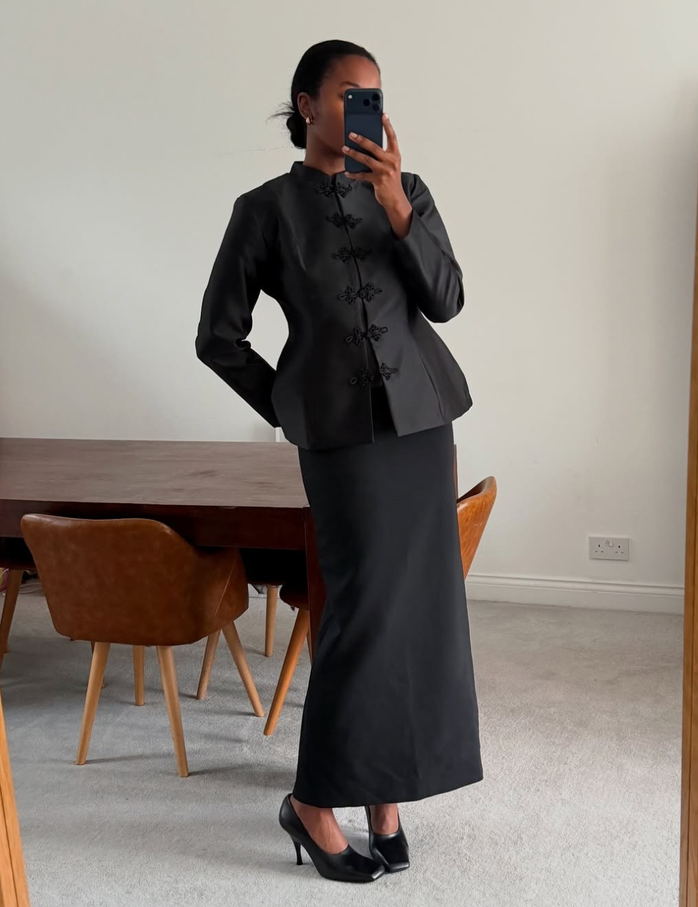 Modelo posa de pé com postura elegante usando vamp pumps pretas de salto fino, saia longa reta e blazer estruturado preto.