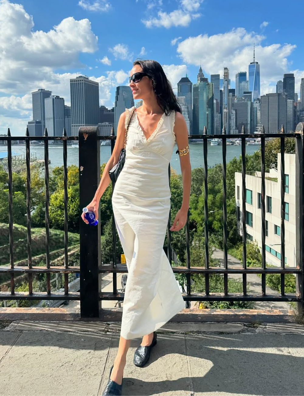 Modelo posa em vestido longo branco de tecido leve com decote em V e babados, combinando vamp pumps pretas de couro.