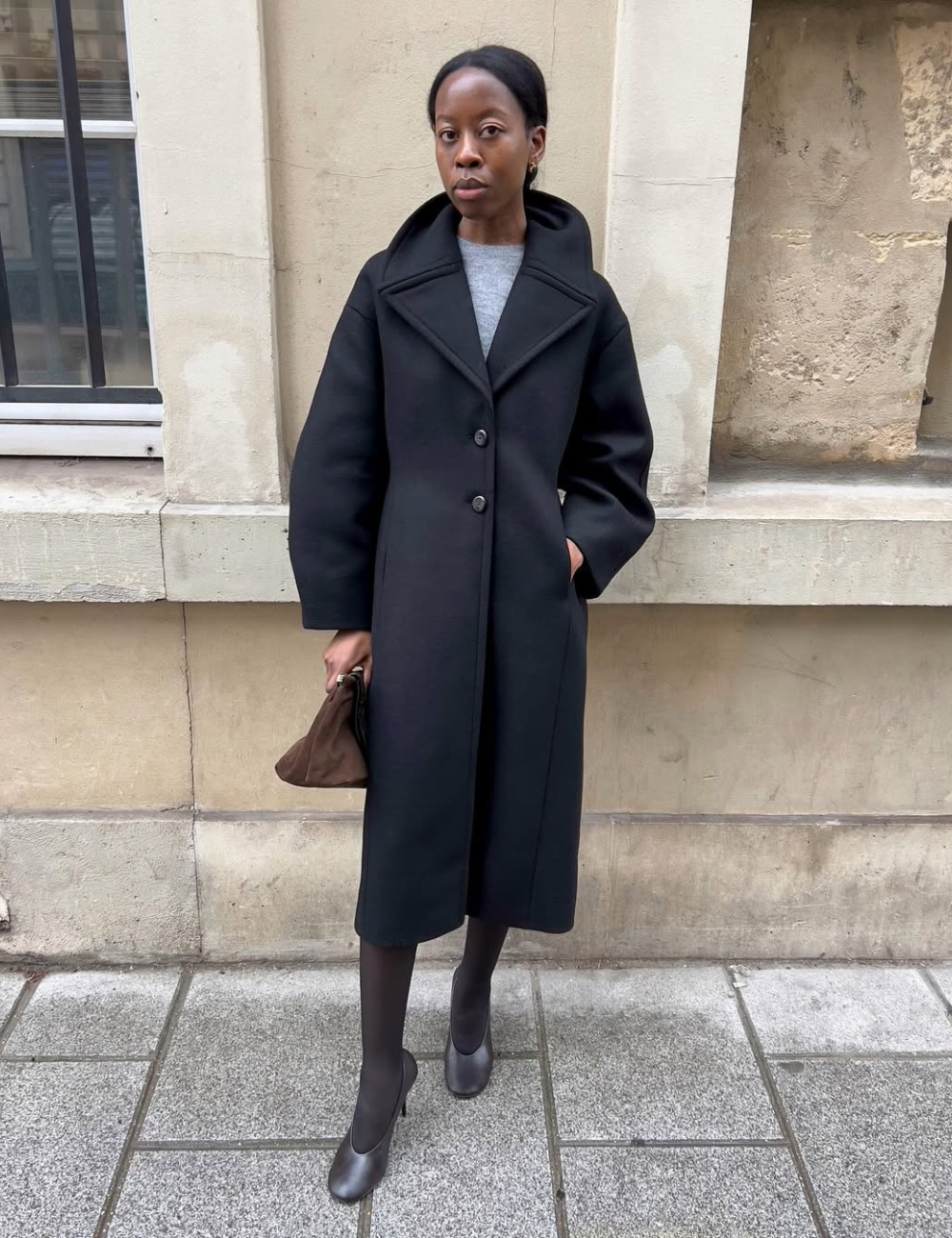 Modelo posa com casaco preto alongado de lã, blusa cinza, meia-calça preta e vamp pumps em couro cinza, em cenário urbano.