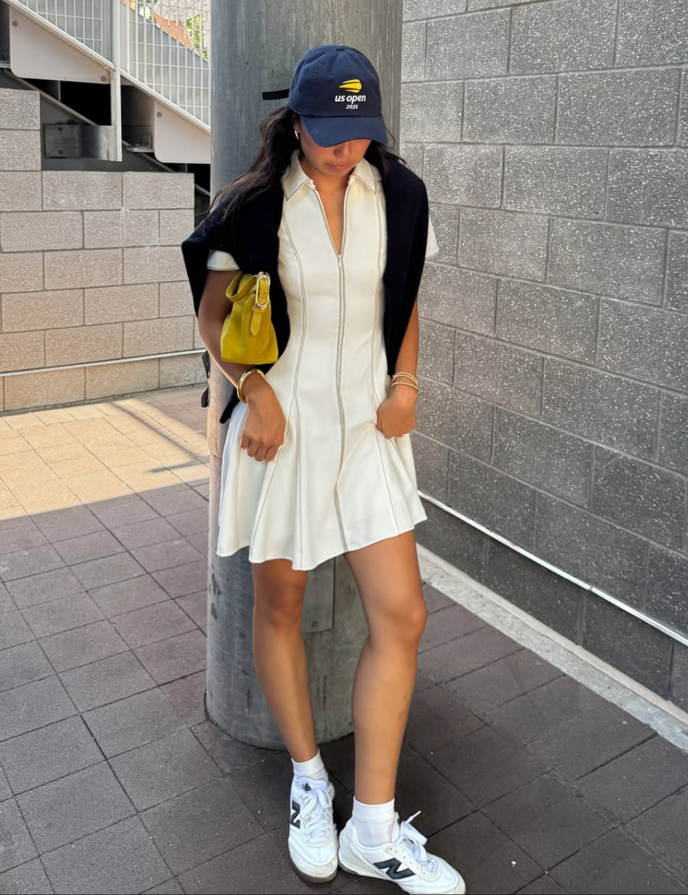 Modelo posa com vestido com tênis branco, boné azul, tricô preto e bolsa amarela em corredor urbano.