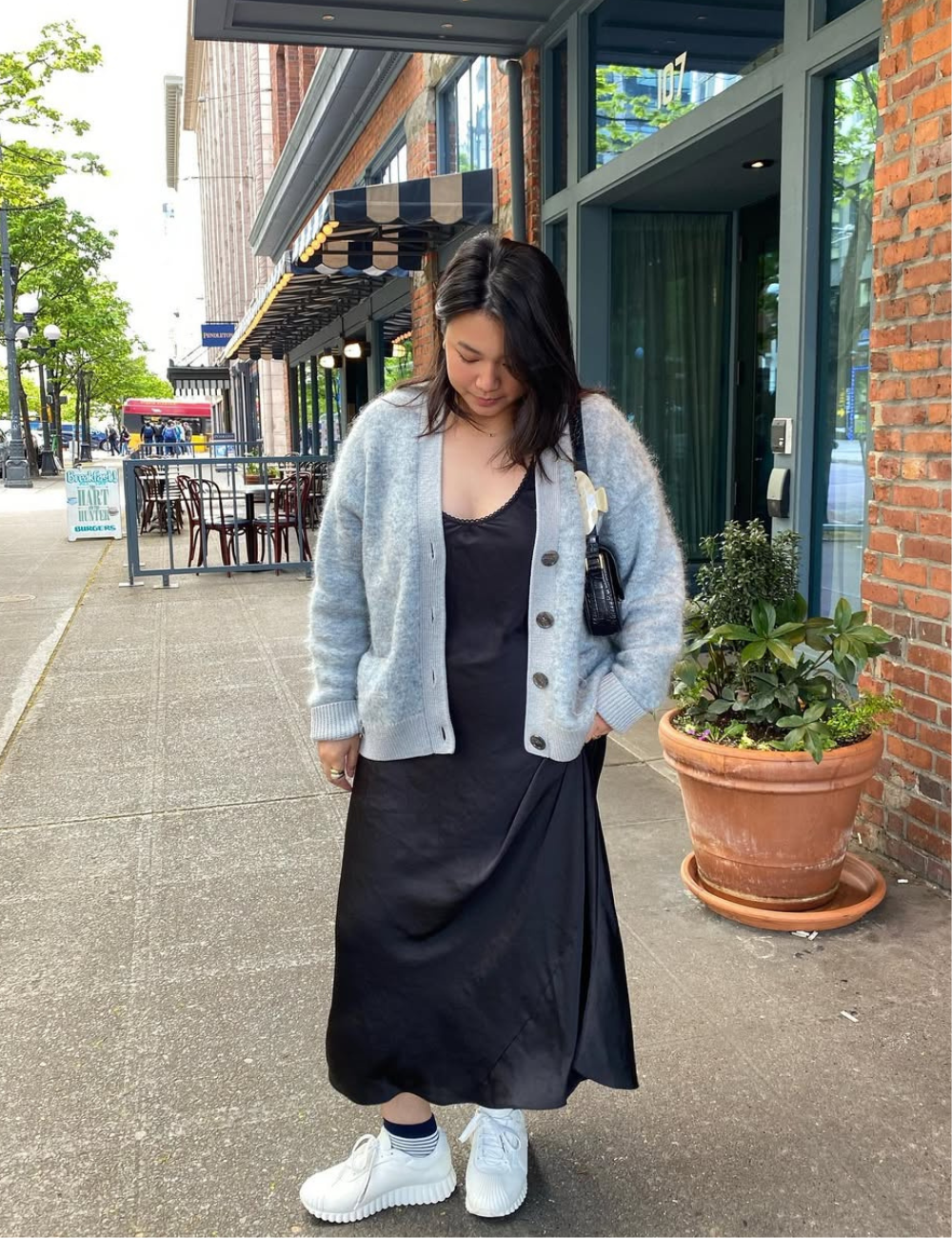 Vestido com tênis branco, modelo posa na calçada com slip dress preto acetinado e cardigan cinza felpudo, em clima casual.