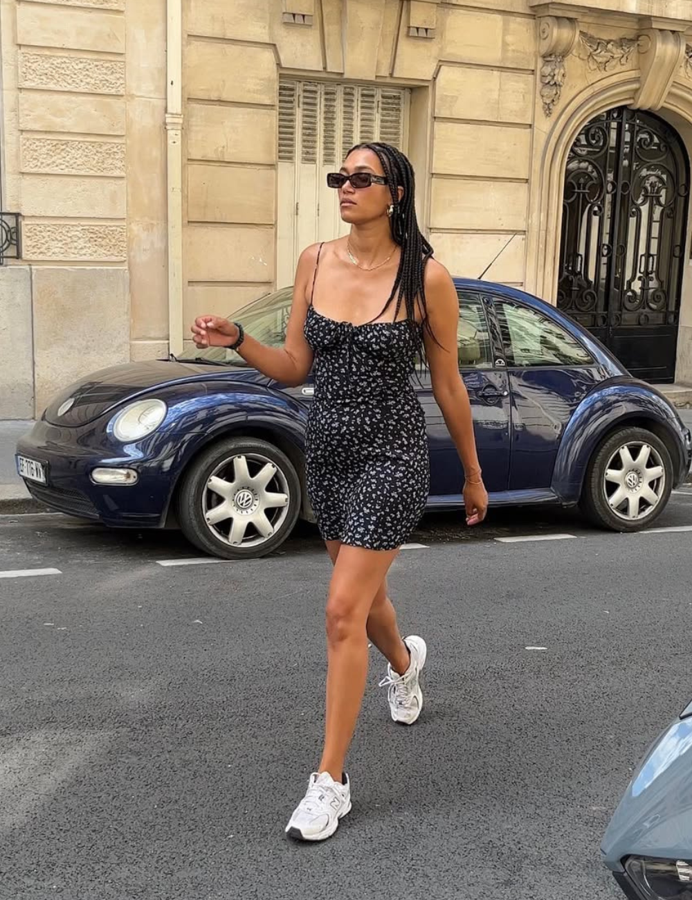 Modelo caminha de vestido com tênis branco, peça preta floral justa, óculos escuros e mood urbano elegante.