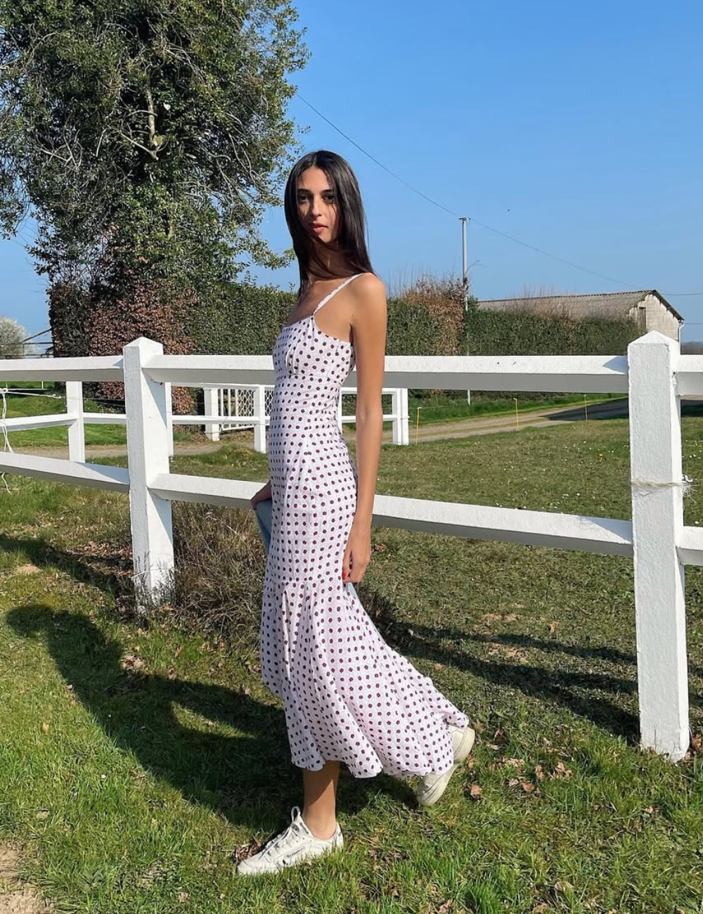 Modelo posa ao ar livre com vestido midi branco estampado e tênis branco, em clima leve e elegante.