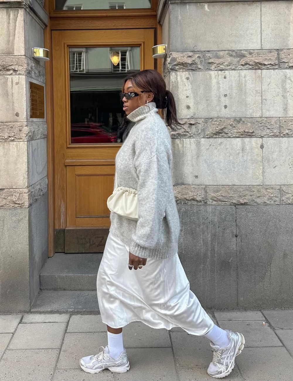 Vestido com tênis branco em cetim e tricô cinza; modelo caminha com bolsa off-white diante de fachada de pedra.