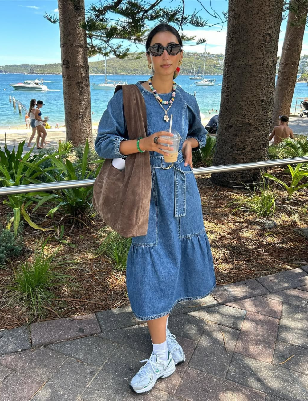 Modelo posa com vestido com tênis branco, jeans midi soltinho, bolsa marrom e óculos escuros à beira-mar serena.
