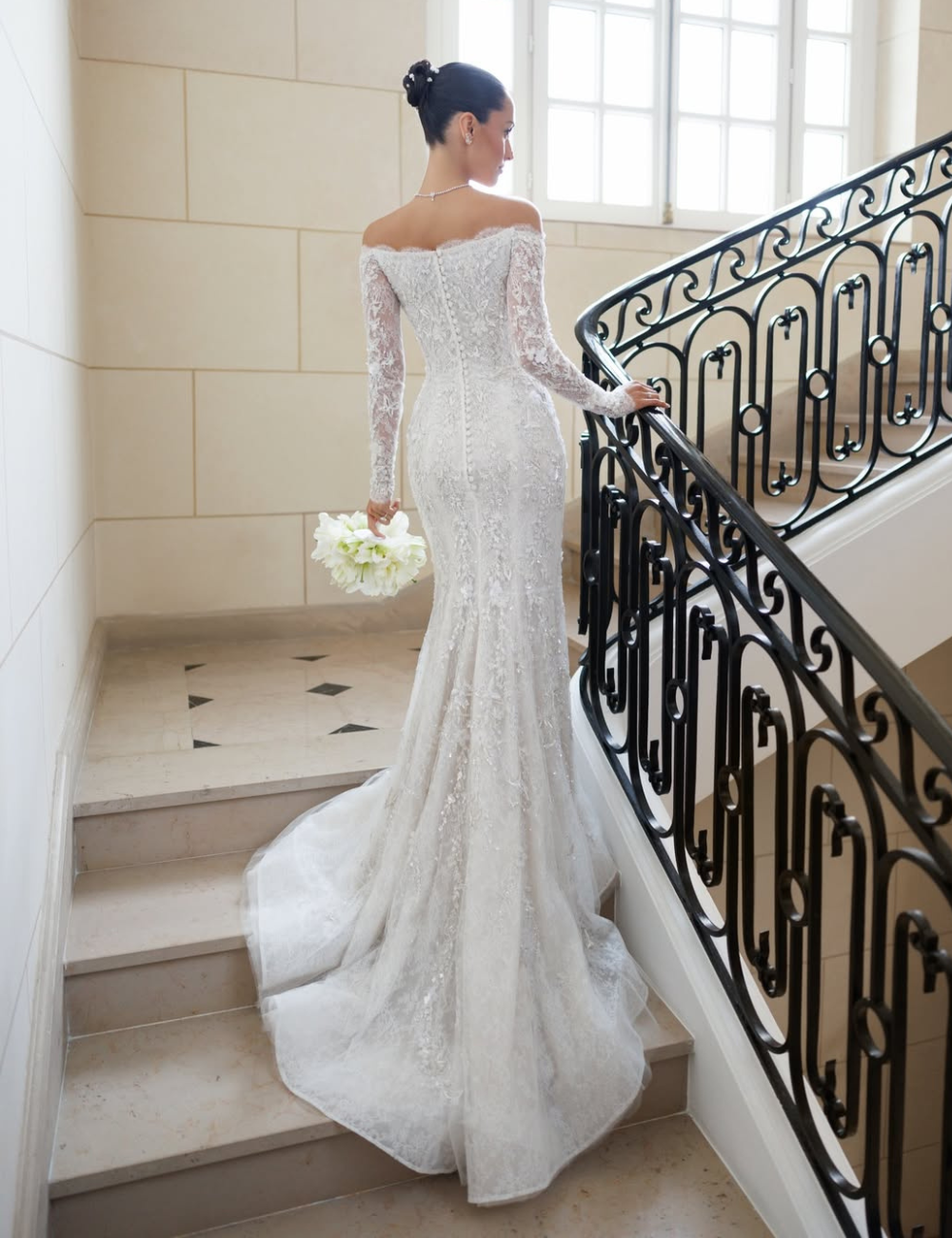 Modelo posa de costas em escada clássica com vestido de noiva da Alexandra Saint Mleux em renda branca.