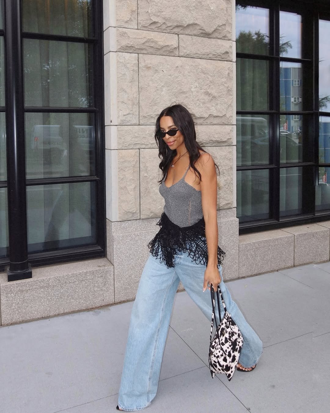 Looks com calça wide leg: modelo posa com jeans claro, top cinza brilhante e franjas pretas em clima urbano elegante.