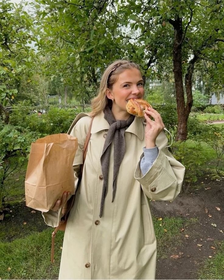 Acessórios para o outono em look bege e marrom: modelo posa no parque com trench coat amplo, lenço e bolsa de couro.