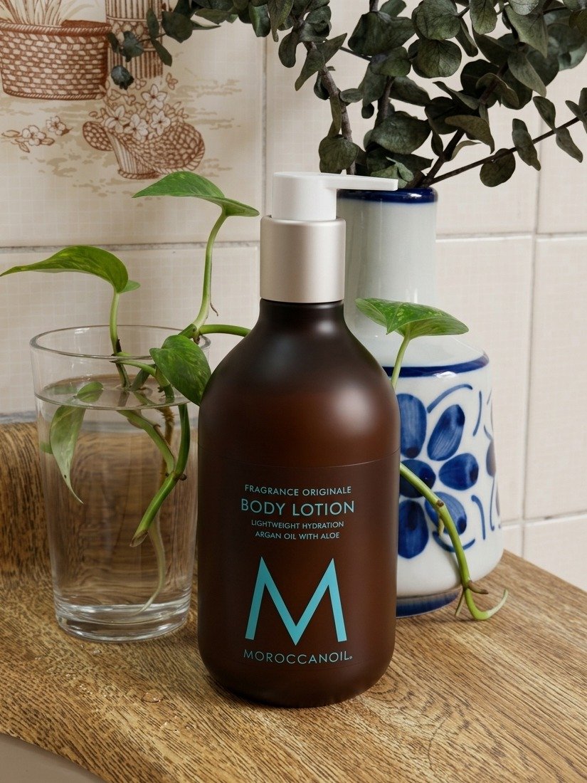 Loção corporal da Moroccanoil em frasco âmbar fosco com pump branco, entre folhagens e cerâmica azul, clima natural;