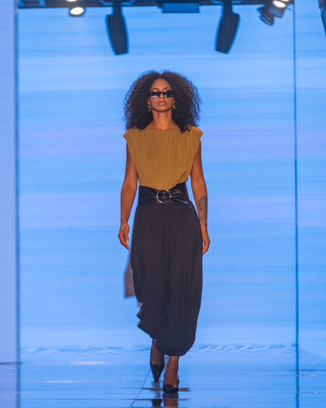 Moda outono inverno 2026: modelo posa na passarela com vestido marrom e preto, cinto largo e clima sofisticado.