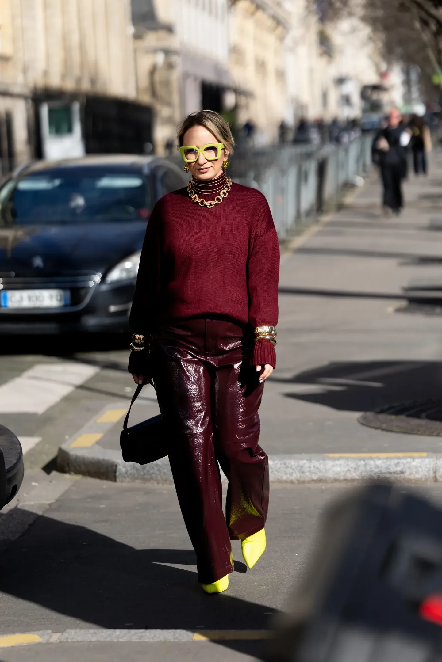 Looks com calça de couro vinho brilhante: modelo posa na rua com tricô amplo, óculos e sapatos neon em clima ousado.