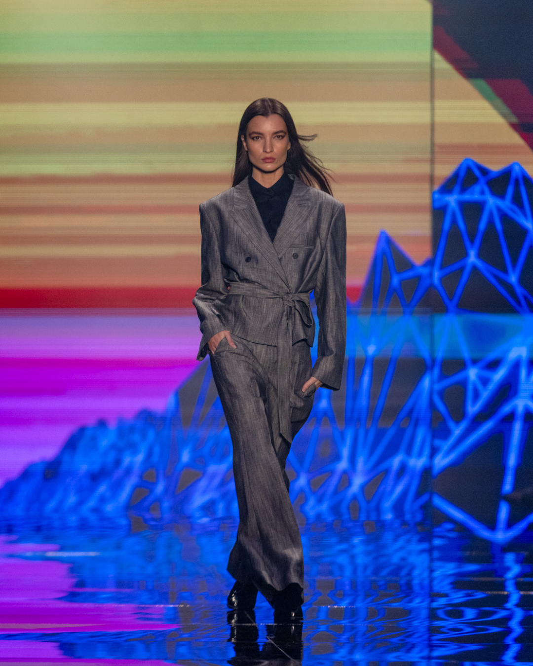 Moda outono inverno 2026: modelo posa com terno cinza amplo e fluido em passarela neon, clima urbano e sofisticado.